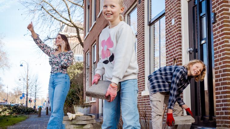 2 kinderen staan met een tegel in hun handen bij een stapel tegels. Op de achtergrond staat een vrouw te bellen