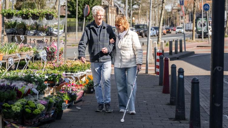 Een vrouw met blindegeleidestok loopt naast een man