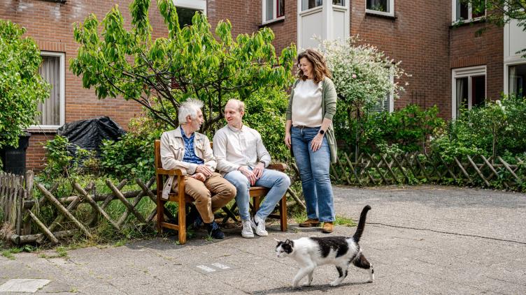 mensen zitten op een bankje voor een appartementencomplex met groene struiken