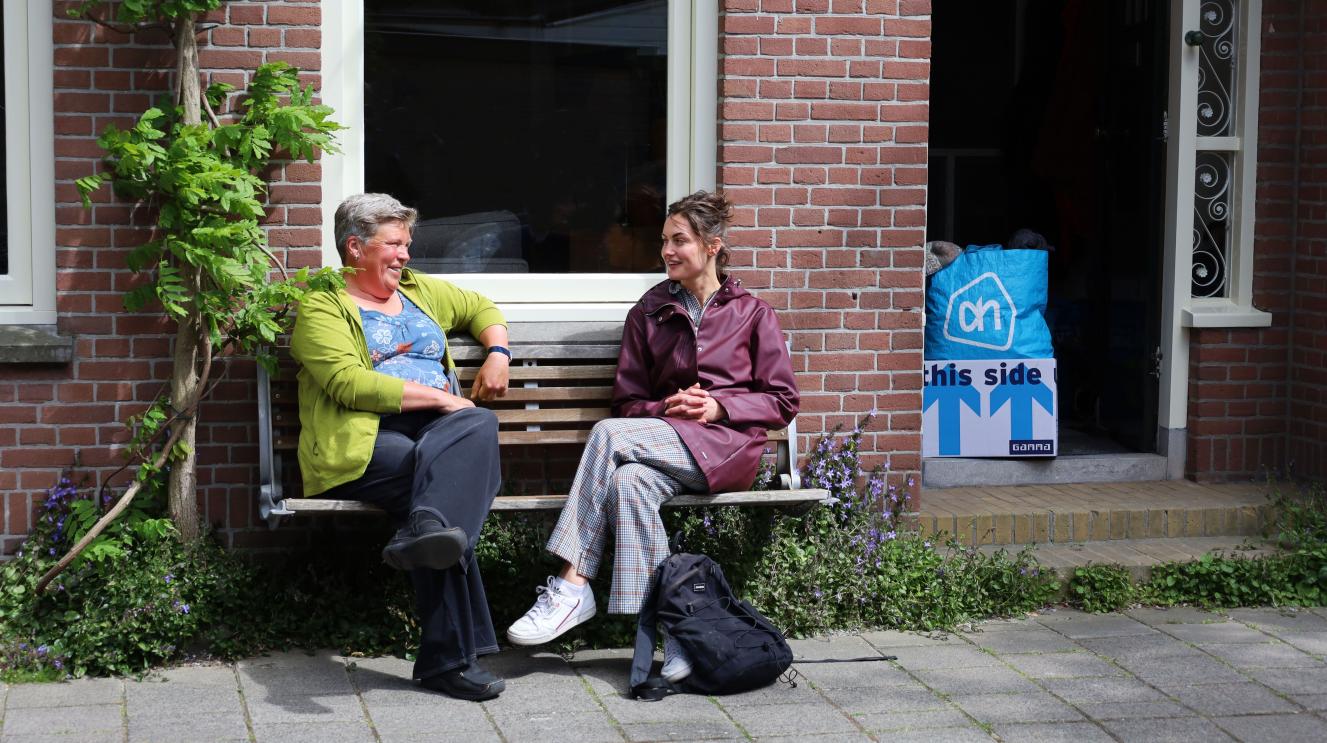 2 vrouwen zitten op een bankje voor een huis. In de deuropening staan verhuisspullen.