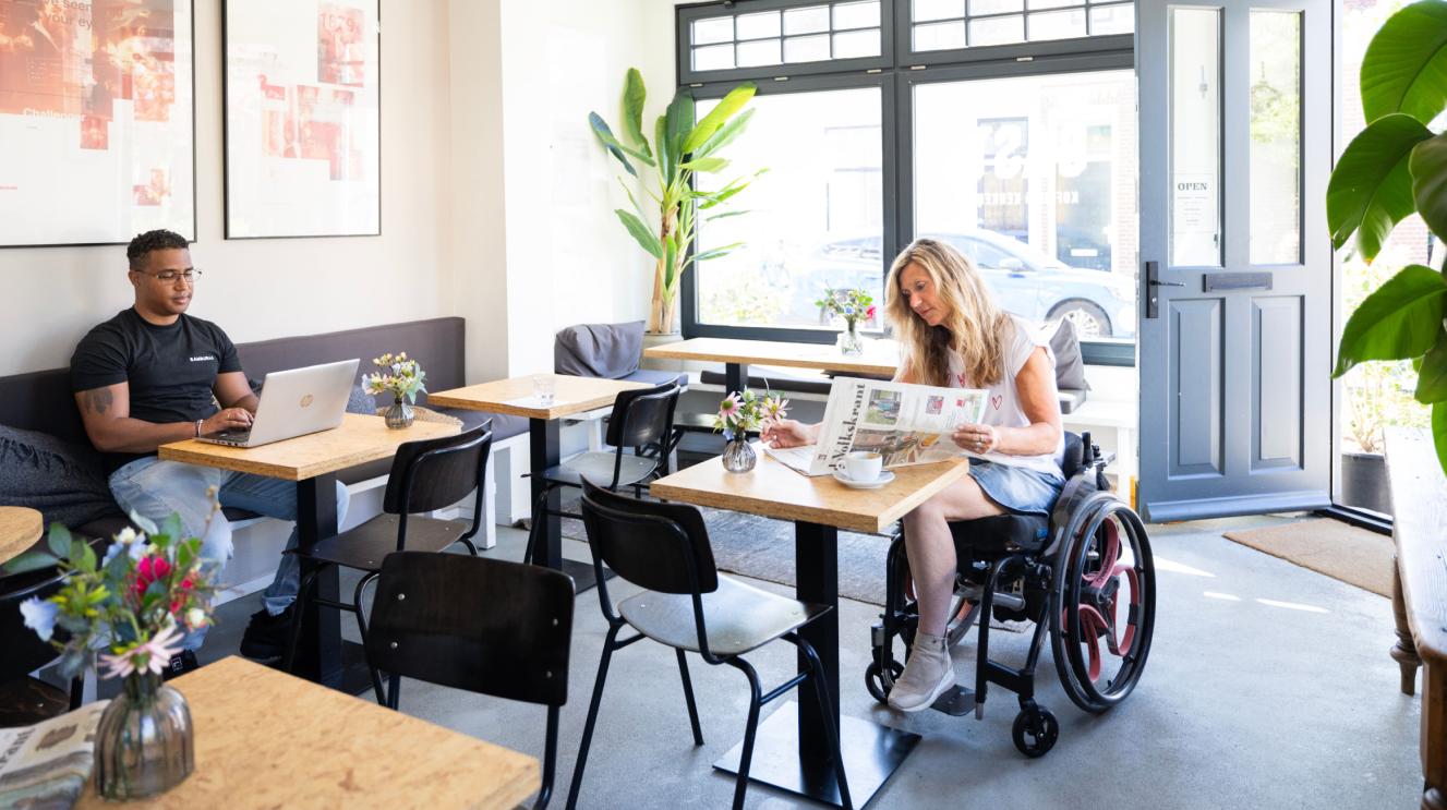 Man met laptop op bank en krantlezende vrouw in rolstoel in café